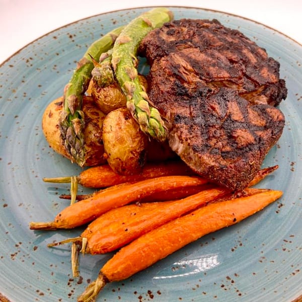 Grilled steak with asparagus, roasted potatoes, and carrots on a blue speckled plate.