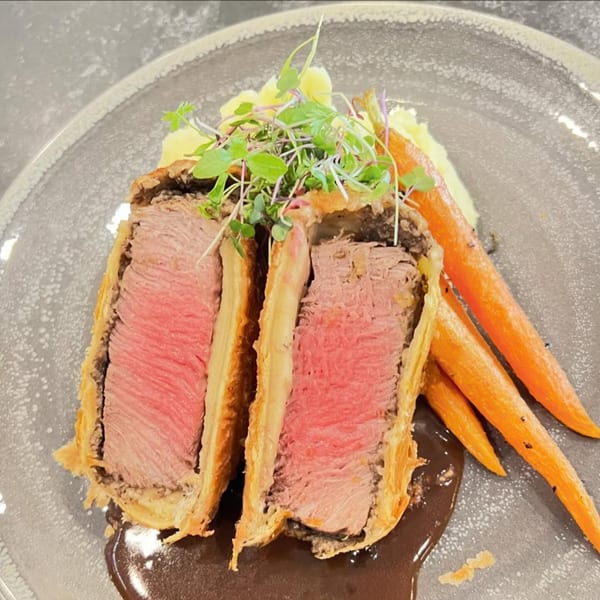 Beef Wellington slices with mashed potatoes, roasted carrots, and gravy on a gray plate.