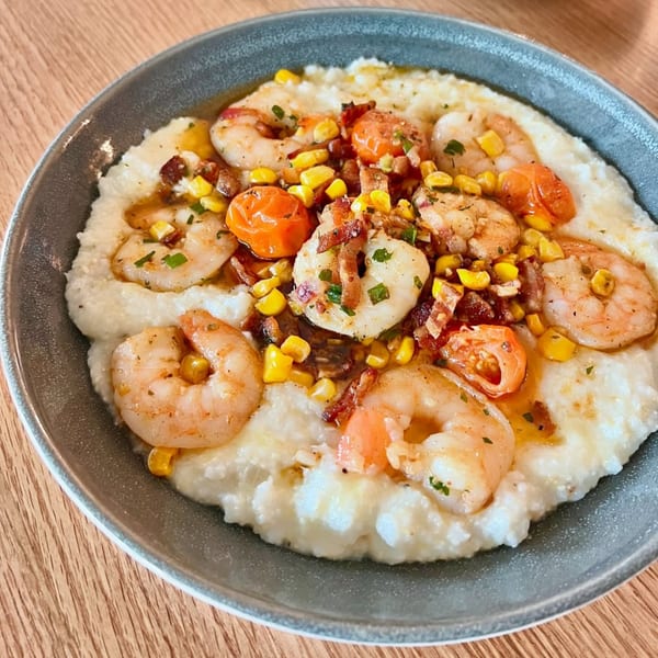 Shrimp and grits with corn, tomatoes, and bacon in a gray bowl on a wooden table.