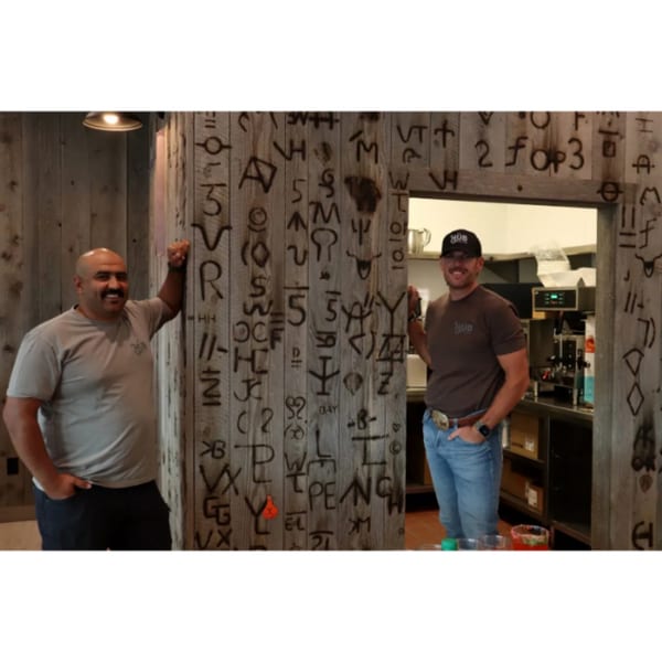Two men in Hub shirts stand by a wooden wall with branded symbols, one in a doorway.