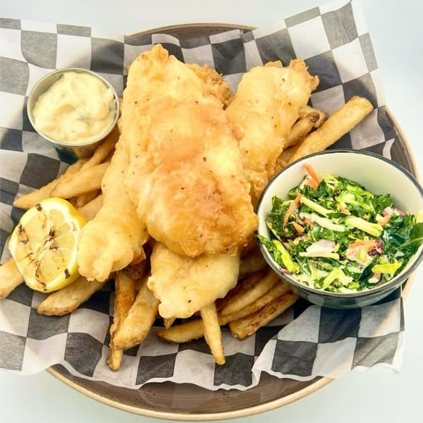 Golden-brown fish and chips with coleslaw, tartar sauce, and a grilled lemon in a basket.