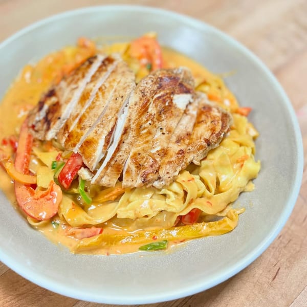 Grilled chicken breast slices over creamy pasta with red and orange bell peppers in a gray bowl.